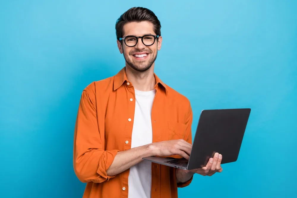 A happy remote worker in a bright orange shirt holding a laptop as he surfs the web learning about business process outsourcing best practices.