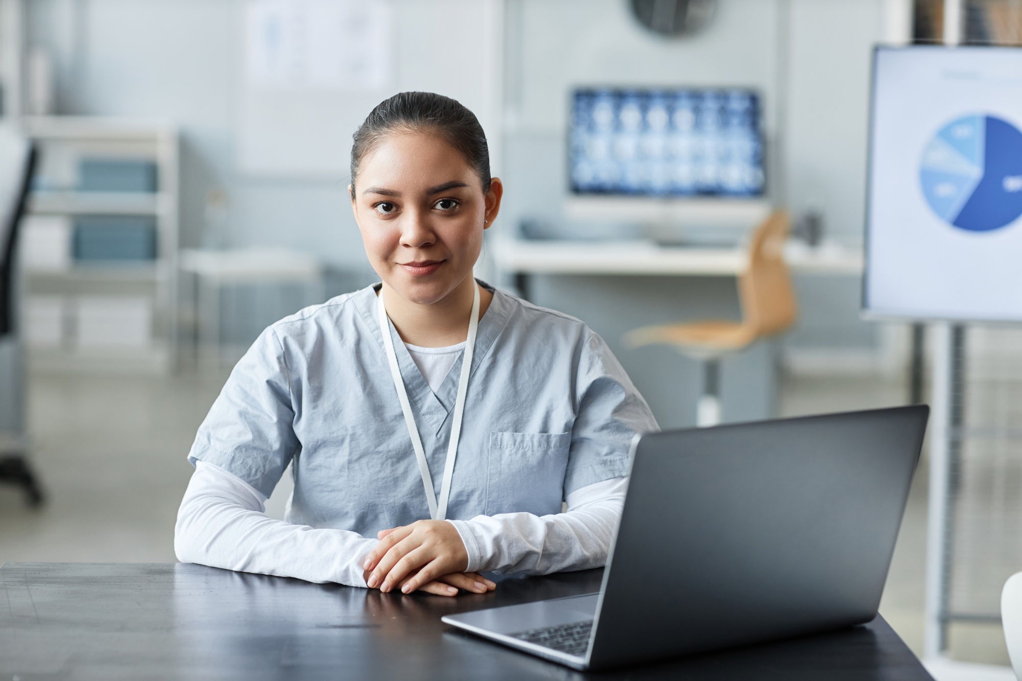 Young pretty female virtual medical assistant poses in front of her laptop in a remote medical setting